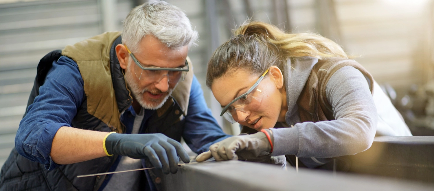 Zwei Personen arbeiten gemeinsam konzentriert an einem Metallstück in einer Werkstatt. Beide tragen Schutzbrillen und Handschuhe. Das Bild vermittelt Teamarbeit und praktisches Arbeiten in einer industriellen oder handwerklichen Umgebung.