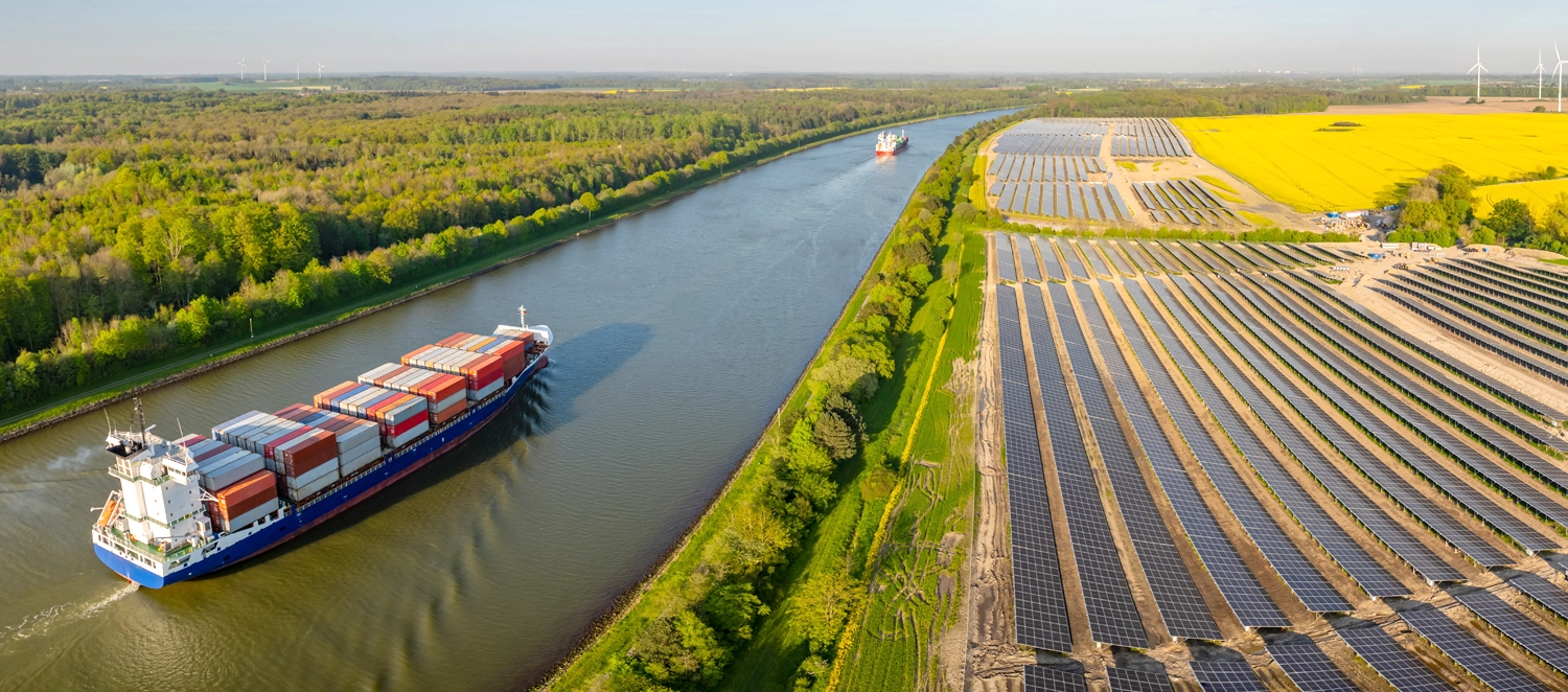 Landschaftsansicht mit einem Fluss, auf dem ein Frachtschiff unterwegs ist, flankiert von großen Solarenergie-Anlagen auf der rechten Seite und landwirtschaftlichen Feldern im Hintergrund. Weiter hinten sind Windkraftanlagen und blühende Rapsfelder zu sehen. Das Bild symbolisiert nachhaltige Energiequellen und umweltfreundlichen Transport.