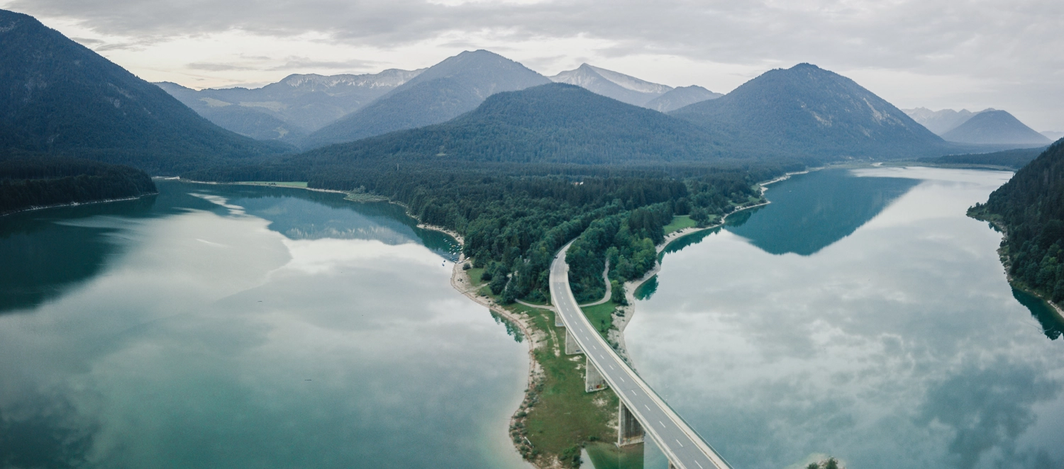 Eine Straße führt über eine lange Brücke, die einen ruhigen, spiegelglatten See überquert, umgeben von bewaldeten Bergen. Die Landschaft ist in Nebel gehüllt, was eine ruhige und friedliche Atmosphäre schafft. Das Bild vermittelt Naturverbundenheit und zeigt eine harmonische Infrastruktur inmitten einer idyllischen Berglandschaft.